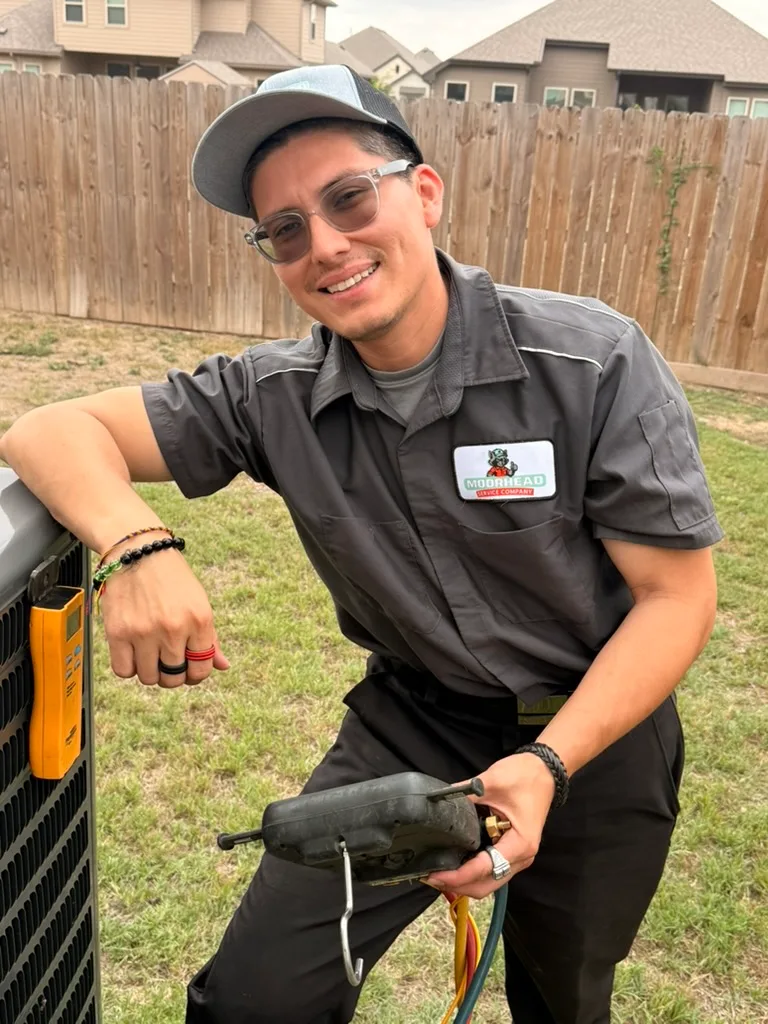 A smiling Moorhead Service Company technician kneeling beside an outdoor AC unit at a Texas home.